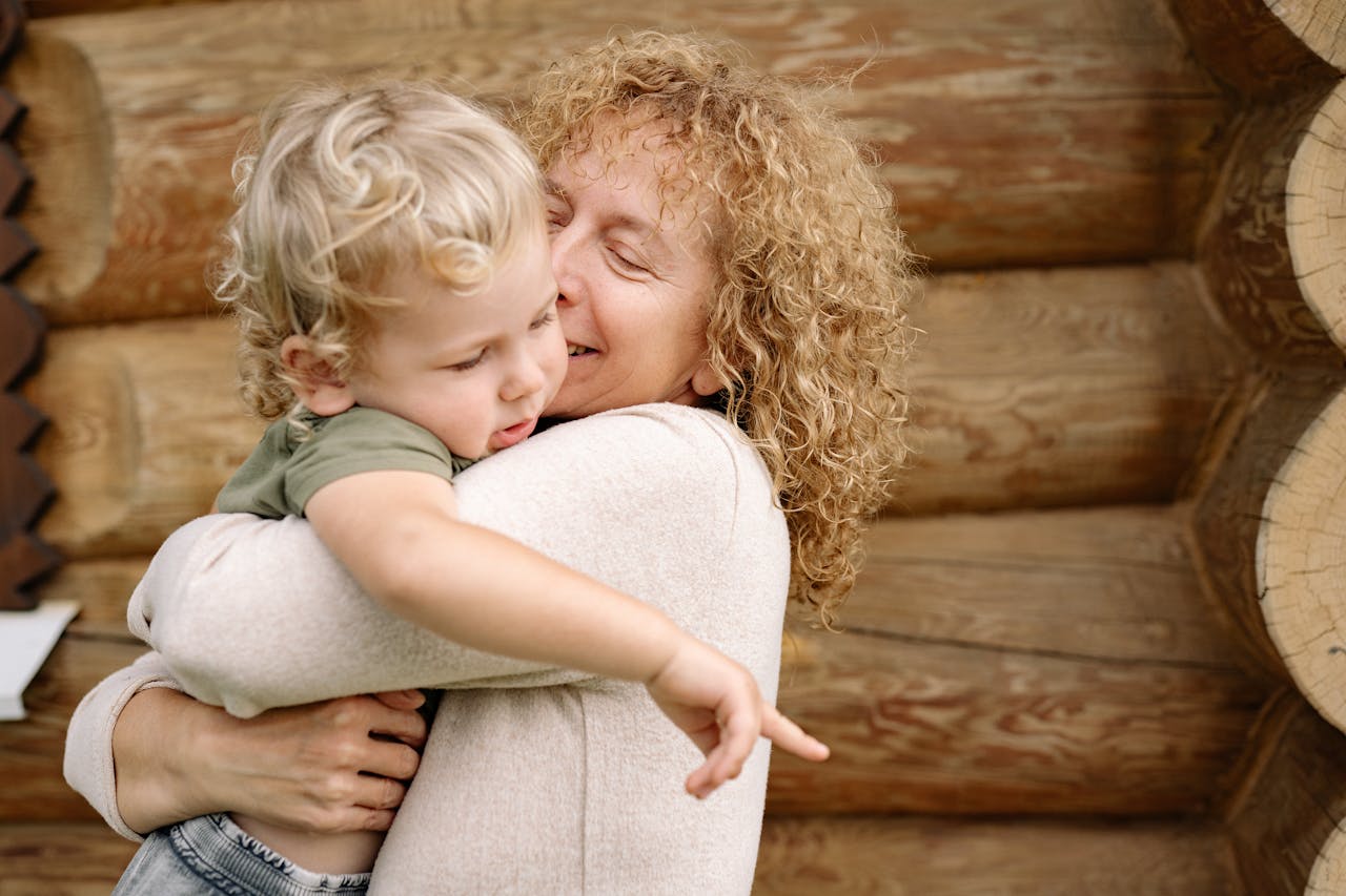 A cheerful grandmother embraces her smiling grandson against a rustic log cabin.